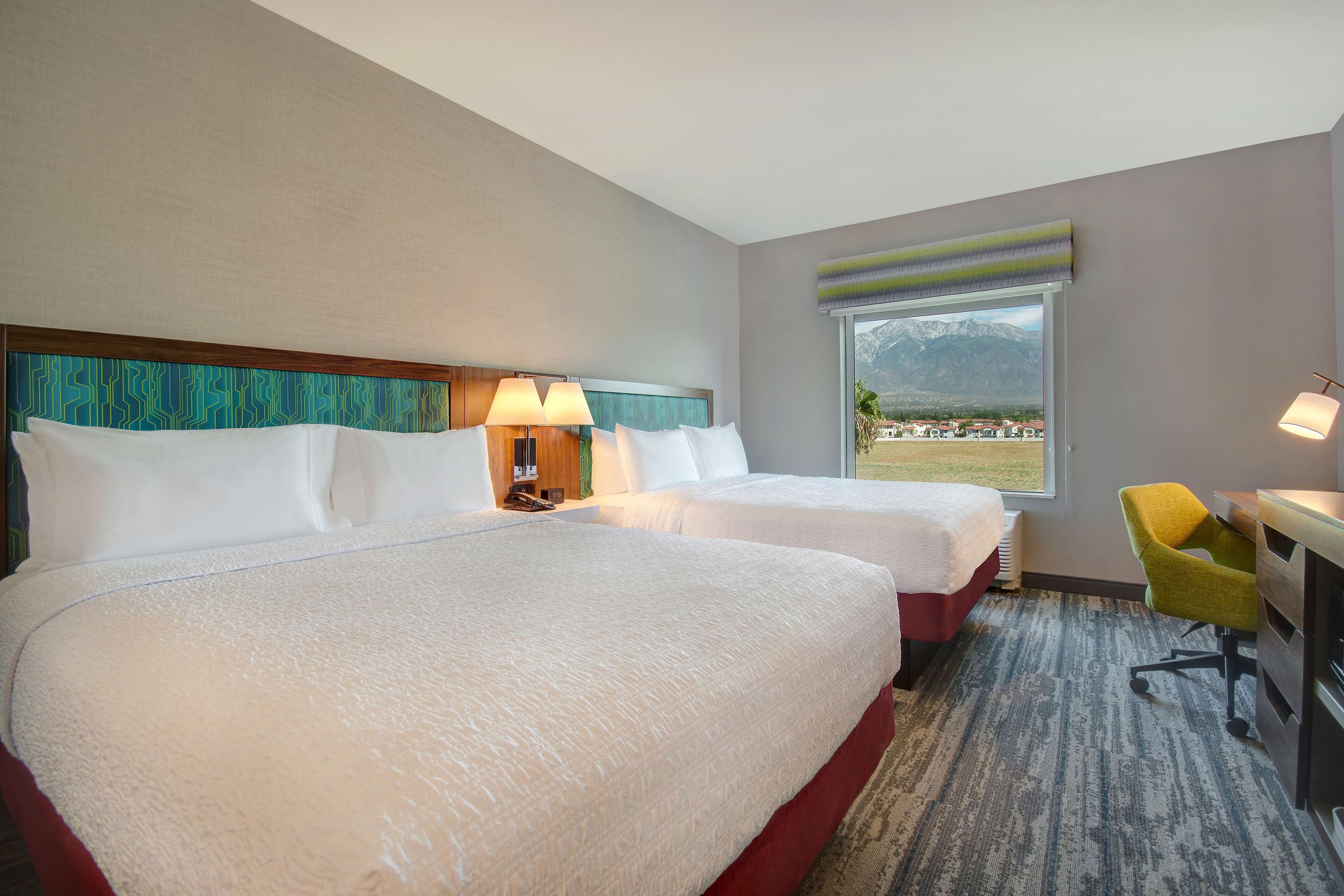 Une chambre d’hôtel avec deux lits, un bureau, une chaise et une grande fenêtre donnant sur la montagne.
