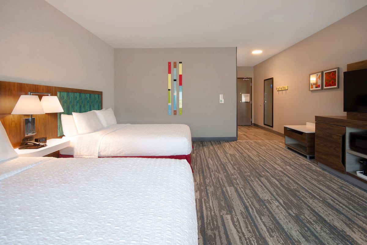 A hotel room with two beds, a desk with a lamp, a TV, and a wall-mounted artwork.