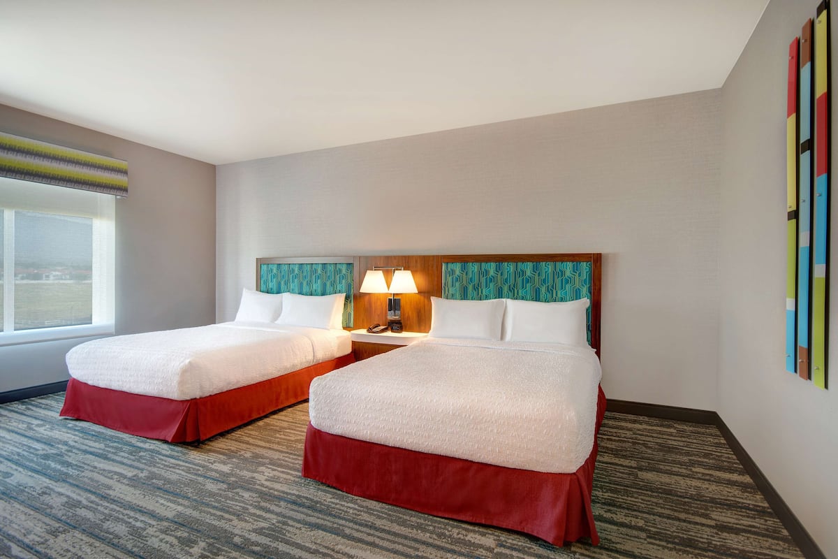 A hotel room with two beds, a window, a lamp, and a wall with colorful artwork.