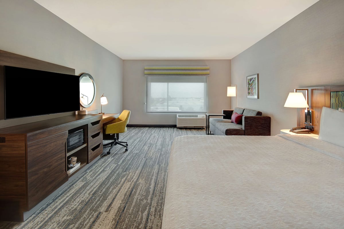 A hotel room with a bed, a desk with a chair, a television, a window with blinds, and a lamp.