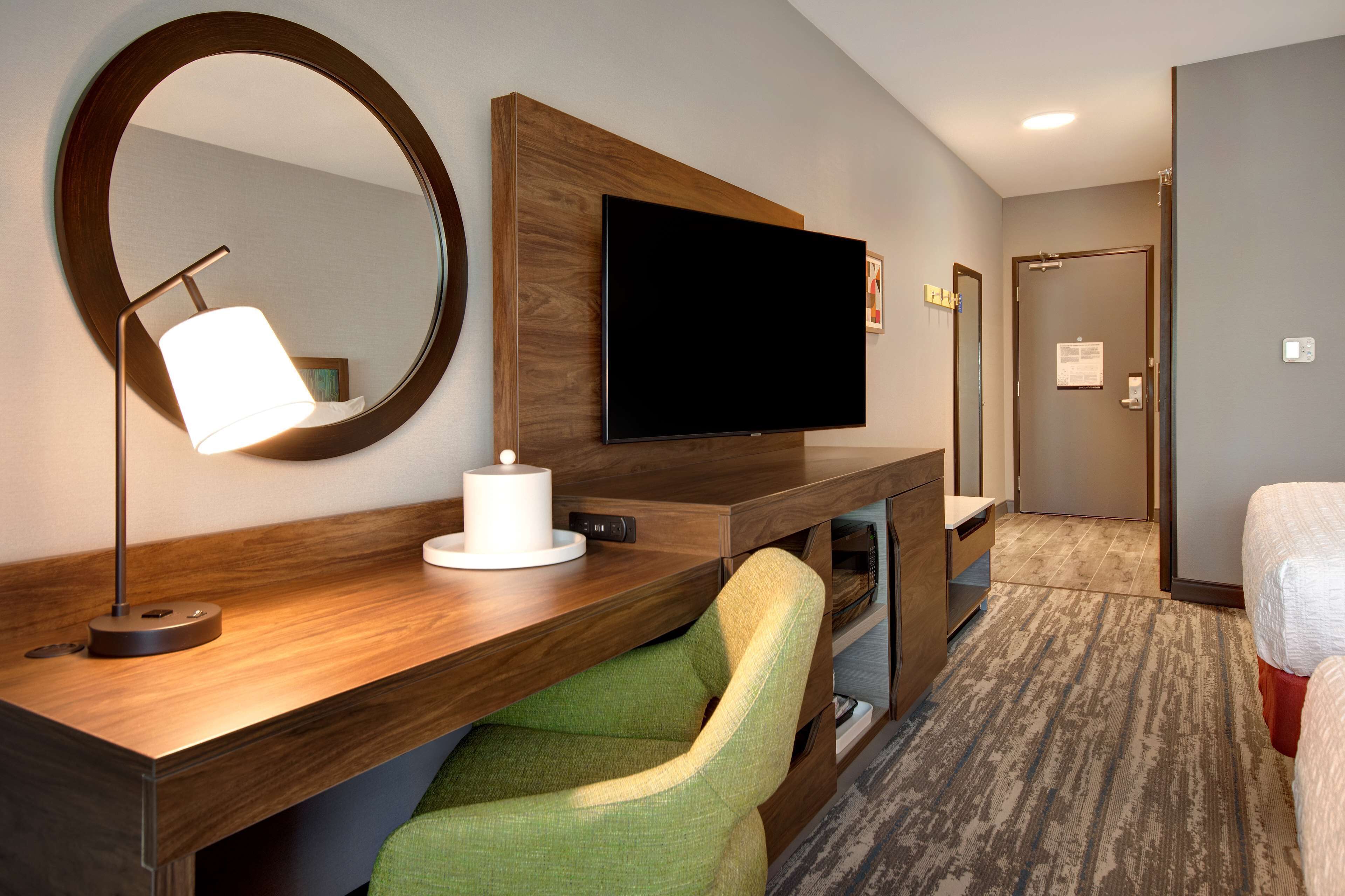 Une chambre d’hôtel avec un bureau en bois, un téléviseur à écran plat, une chaise verte, un miroir rond et un lit.