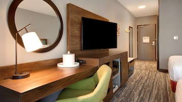 Une chambre d’hôtel avec un bureau en bois, un téléviseur à écran plat, une chaise verte, un miroir rond et un lit.