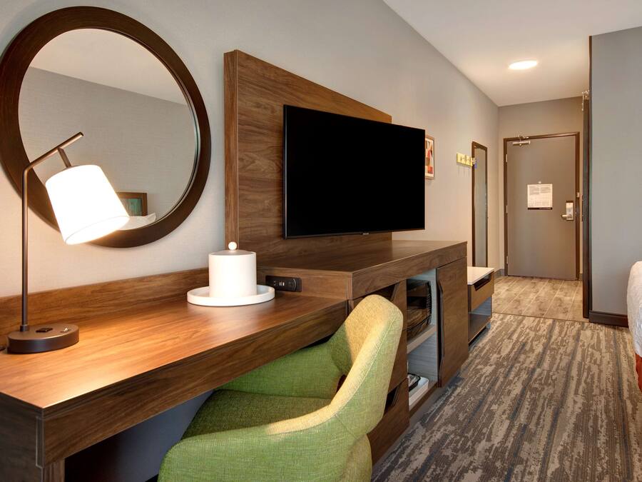 Une chambre d’hôtel avec un bureau en bois, un téléviseur à écran plat, une chaise verte, un miroir rond et un lit.