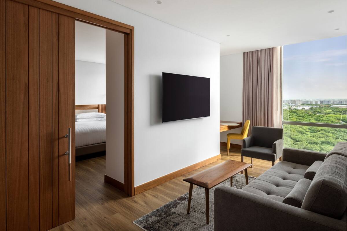 Une chambre d’hôtel moderne dotée d’une armoire en bois, d’un lit, d’un téléviseur à écran plat, d’un canapé, d’un bureau et offrant une vue sur la verdure et les bâtiments.