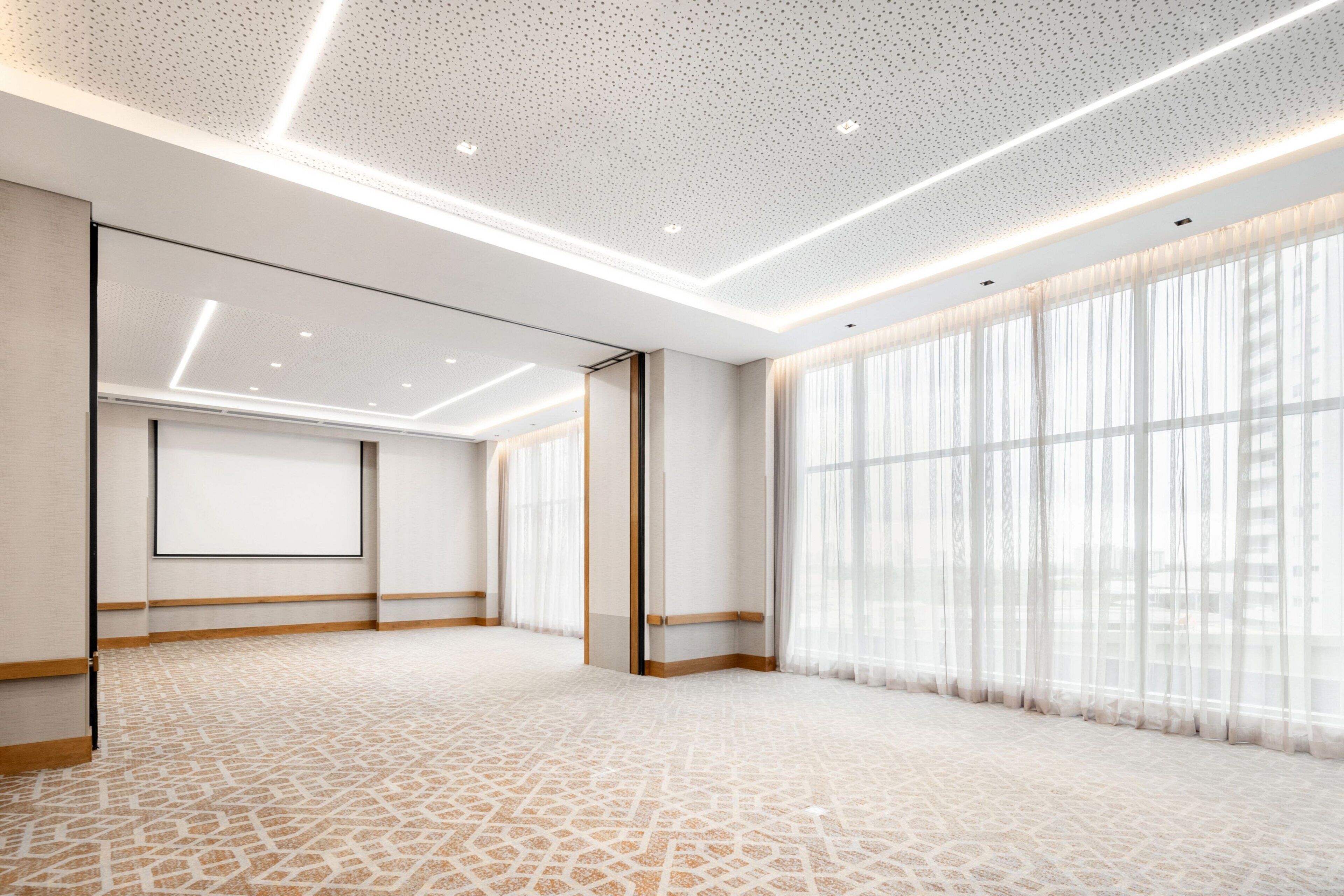 A spacious hotel room with a patterned carpet, large windows with sheer curtains, and a modern ceiling design.