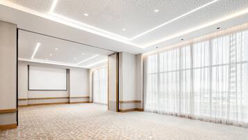 A spacious hotel room with a patterned carpet, large windows with sheer curtains, and a modern ceiling design.