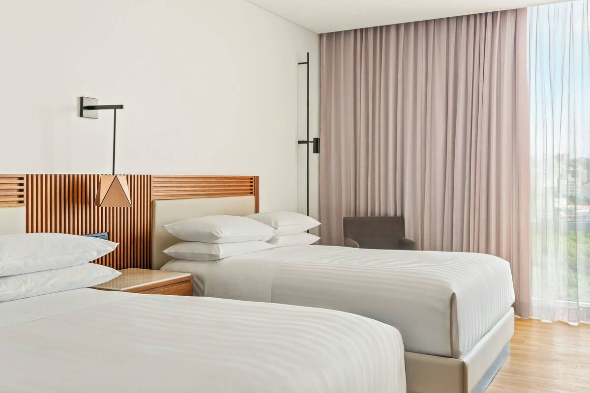 Une chambre d’hôtel avec deux lits, une tête de lit en bois, une table de chevet, une chaise et une grande fenêtre avec des rideaux.