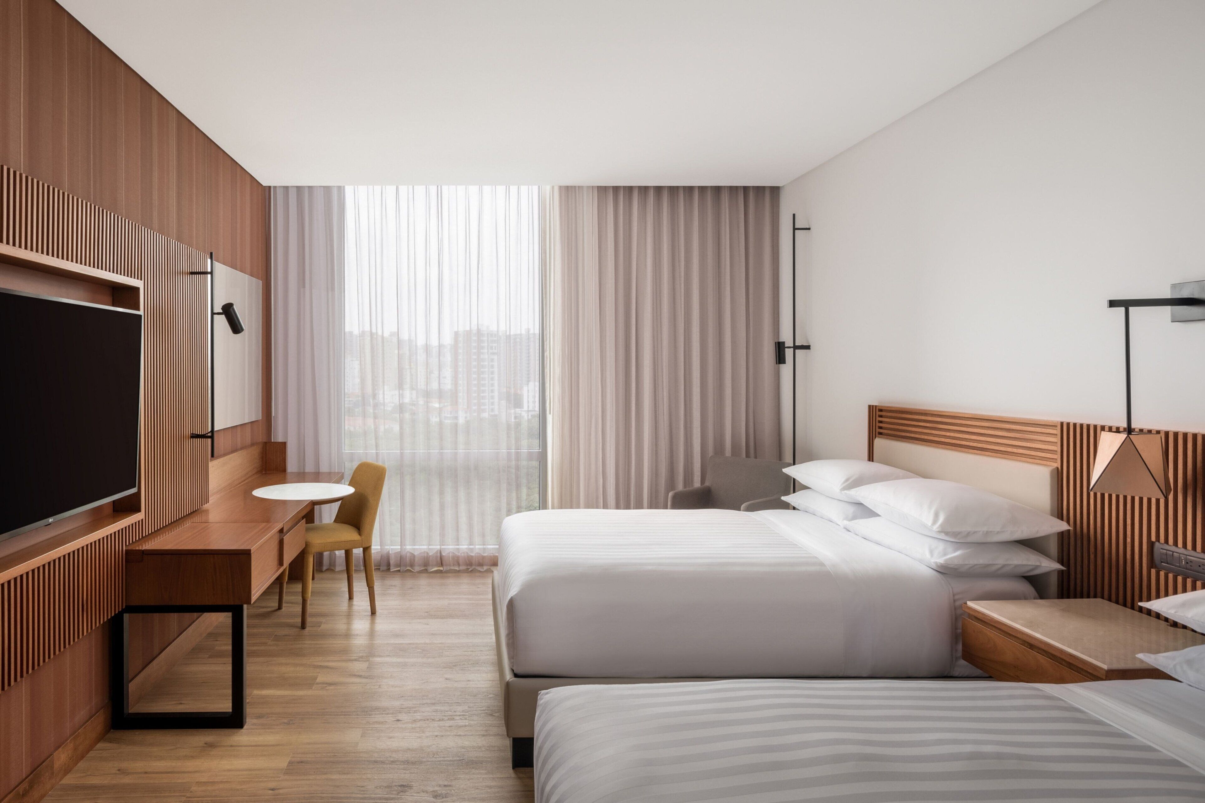 A hotel room with a large bed, a smaller bed, a desk with a chair, a TV, and a window with curtains.