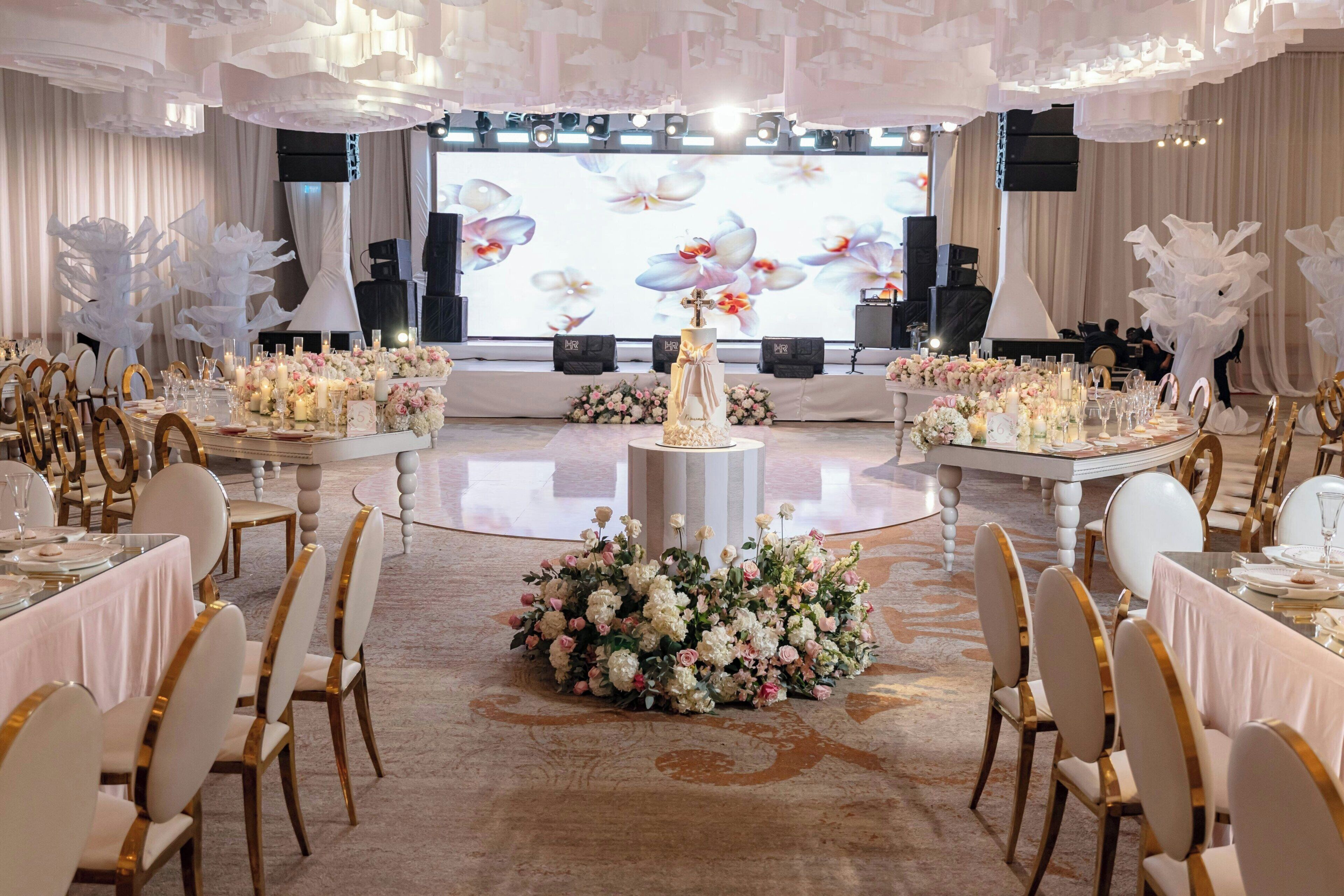 A banquet hall with round tables, chairs, floral centerpieces, and a stage with a floral backdrop.