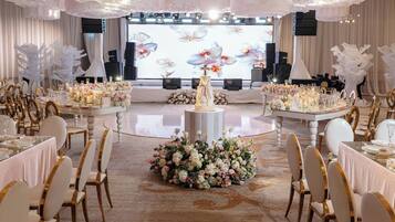A banquet hall with round tables, chairs, floral centerpieces, and a stage with a floral backdrop.