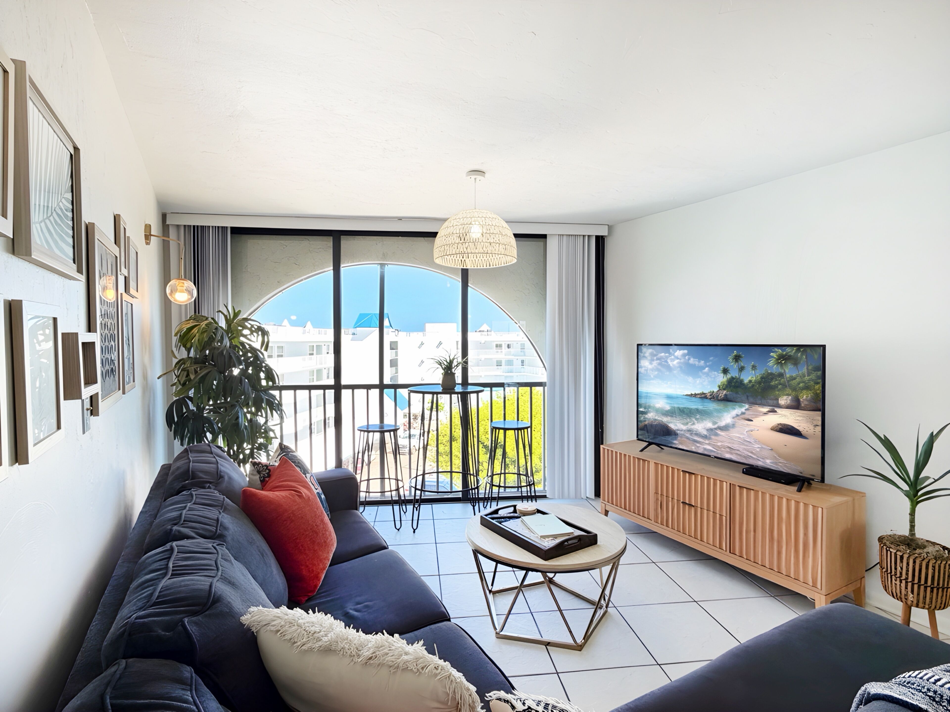 A modern living room with a large TV, comfortable seating, a balcony with a view, and a dining area.