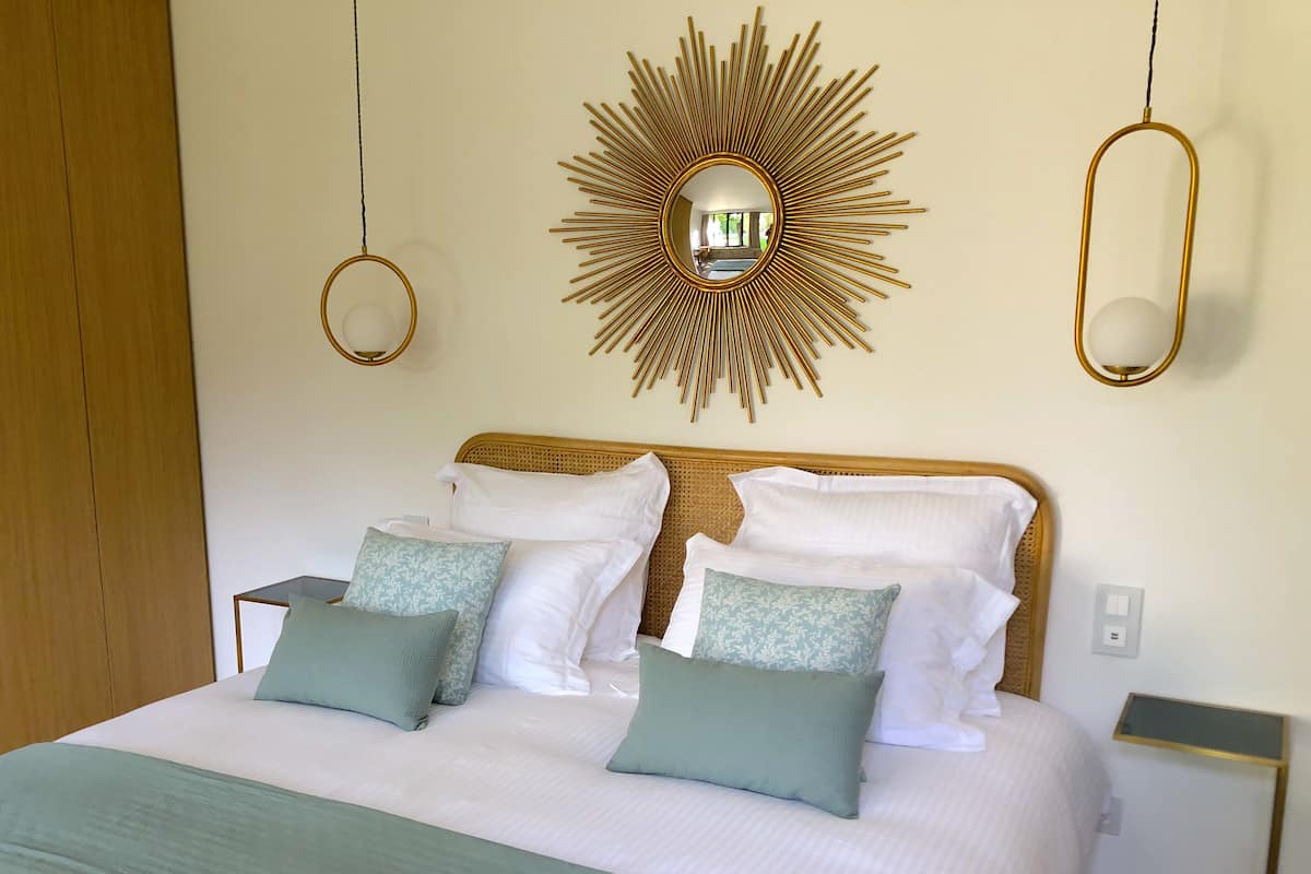 Ein ordentlich bezogenes Bett mit weißen und grünen Kissen, einem Spiegel mit Sonnenstrahlendesign und zwei Pendelleuchten.