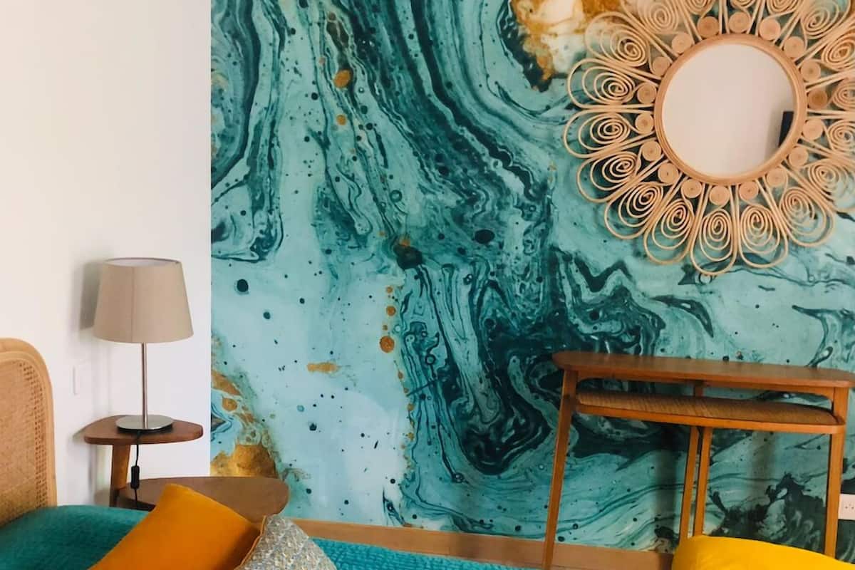 Ein Zimmer mit petrolfarbener Bettwäsche, einem Holfnachtkästchen, einer Lampe und einer Wand mit Marmordesign.