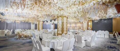 A banquet hall with round tables, white chairs, and a ceiling adorned with floral decorations.