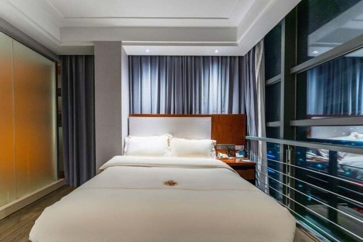 Habitación de hotel con una cama grande, una mesita de noche, un cabecero de madera y vistas de la ciudad a través de ventanales del suelo al techo.