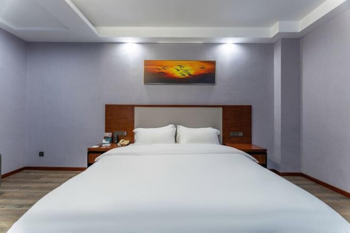 Una habitación de hotel con una cama grande, cabecero de madera, dos mesitas de noche, un cuadro en la pared y una lámpara en el techo.