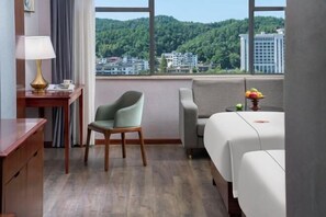 Habitación de hotel con vista a una colina boscosa, un escritorio con lámpara, una silla, un sofá y una cama.