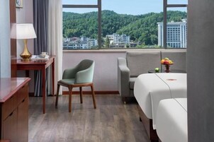 Habitación de hotel con vista a una colina boscosa, un escritorio con lámpara, una silla, un sofá y una cama.