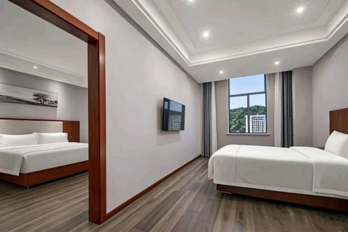 Habitación de hotel con una cama grande, una cama más pequeña, un televisor de pantalla plana, un cuadro en la pared y una ventana que ofrece vistas a árboles y edificios.