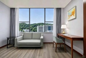 A hotel room with a sofa, a chair, a desk, and a large window offering a view of a cityscape and greenery. - King's Park International Hotel (Tonggu)