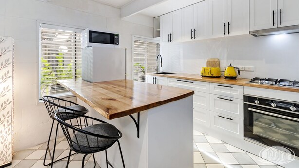 Een moderne keuken met witte kastjes, een houten eiland, twee zwarte stoelen, een magnetron, een waterkoker en een oven.