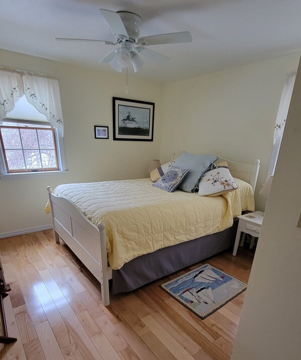 Ein Schlafzimmer mit Bett, Deckenventilator, Fenster mit Vorhängen, einem gerahmten Bild an der Wand und einem kleinen Nachttisch.