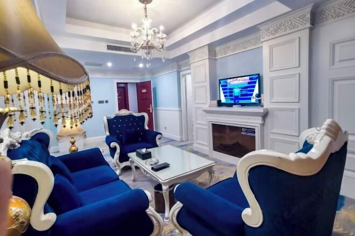 A living room with blue upholstered furniture, a glass coffee table, a chandelier, a fireplace, and a television.