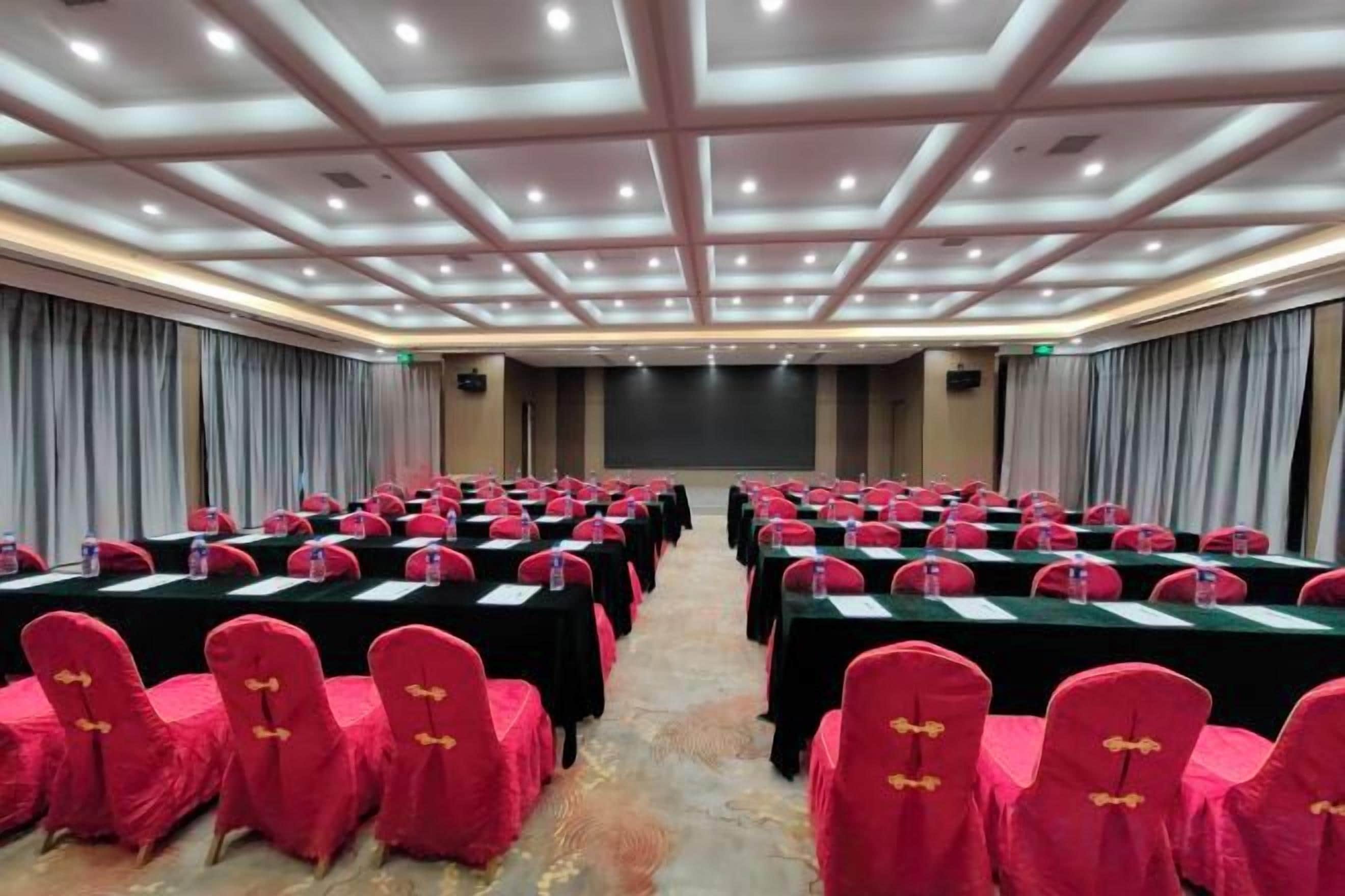 Une salle de conférence avec des rangées de tables et de chaises, un grand écran et une scène.