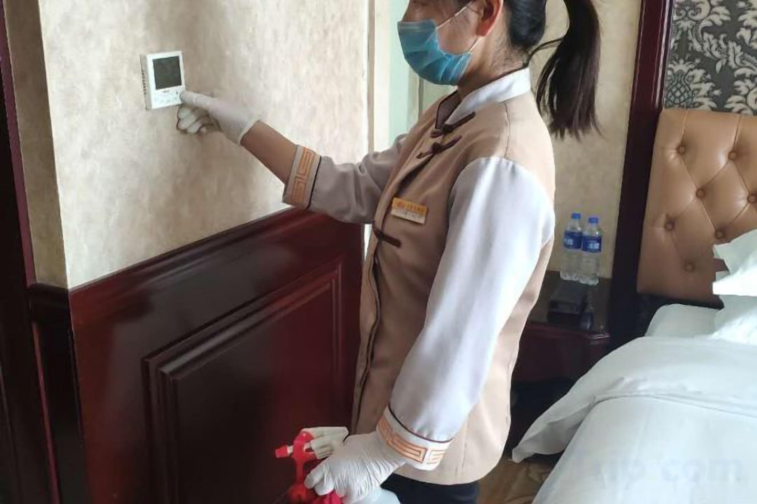 A person in a uniform checking a device on a wall, standing in a room with a bed and a nightstand with bottles.