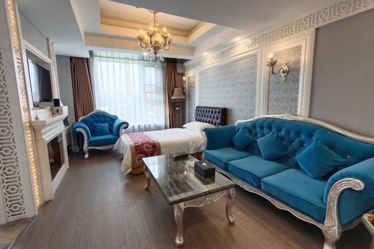 A hotel room with a blue sofa, a white bed, a blue armchair, a glass coffee table, and a chandelier.