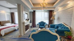 Habitación de hotel con cama, dos sillones, un sofá, una araña y un ventanal con cortinas.