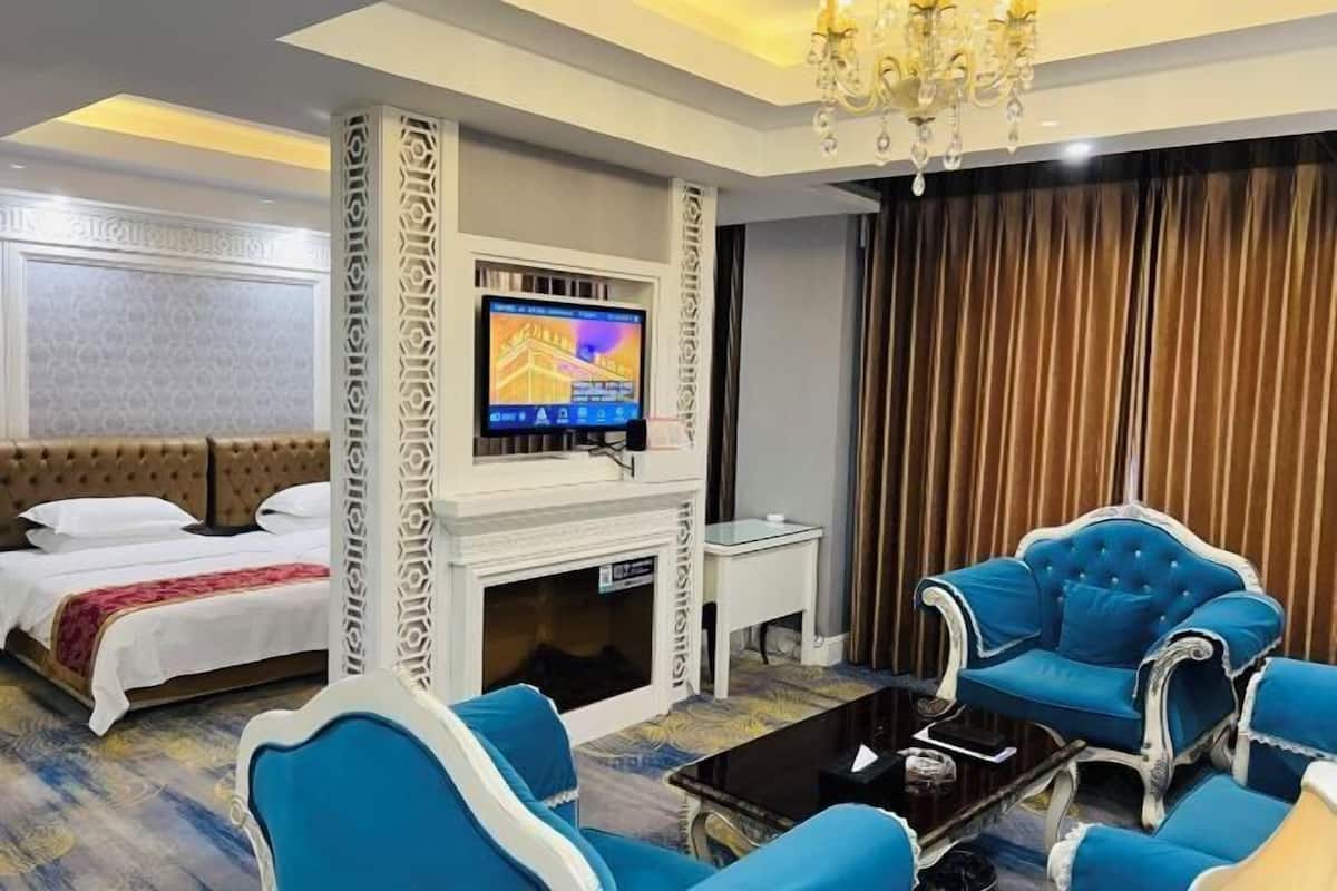 A hotel room with a bed, a TV mounted on a wall, a fireplace, and blue upholstered chairs.