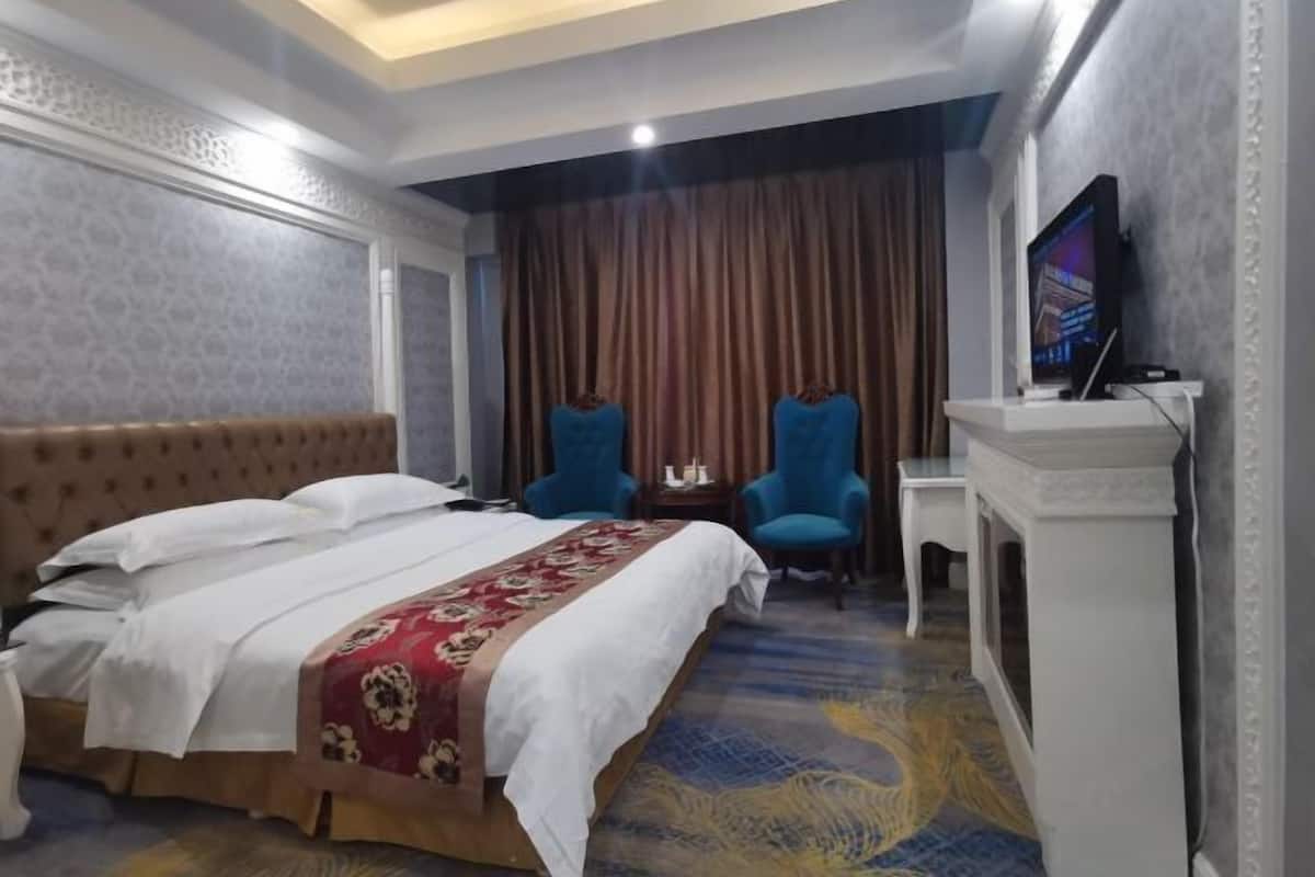 A hotel room with a bed, two blue armchairs, a small table, a TV, and a fireplace.