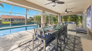 A poolside dining area with a table set for two, chairs, a ceiling fan, and a view of a swimming pool and a house in the background. - Great for kids! Paddle boards, bikes, kayaks & pet friendly! Villa Evelyn's Palms - SWFL Rentals (Cape Coral)