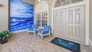 A covered outdoor seating area with two chairs and a potted plant, a large ocean mural, a white door, and a welcome mat. - Great for kids! Paddle boards, bikes, kayaks & pet friendly! Villa Evelyn's Palms - SWFL Rentals (Cape Coral)