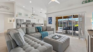A modern living room with a sectional sofa, ottoman, ceiling fans, and a view to a patio with outdoor furniture. - Great for kids! Paddle boards, bikes, kayaks & pet friendly! Villa Evelyn's Palms - SWFL Rentals (Cape Coral)