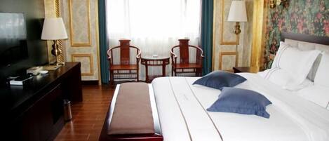 A hotel room with a large bed, a desk with a TV, two chairs, and a small table.