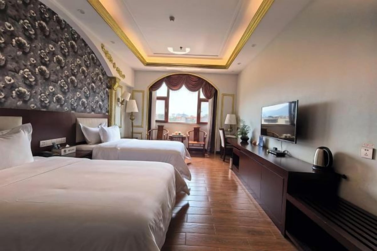 A hotel room with two beds, a television, a desk with a chair, and a view through a window.