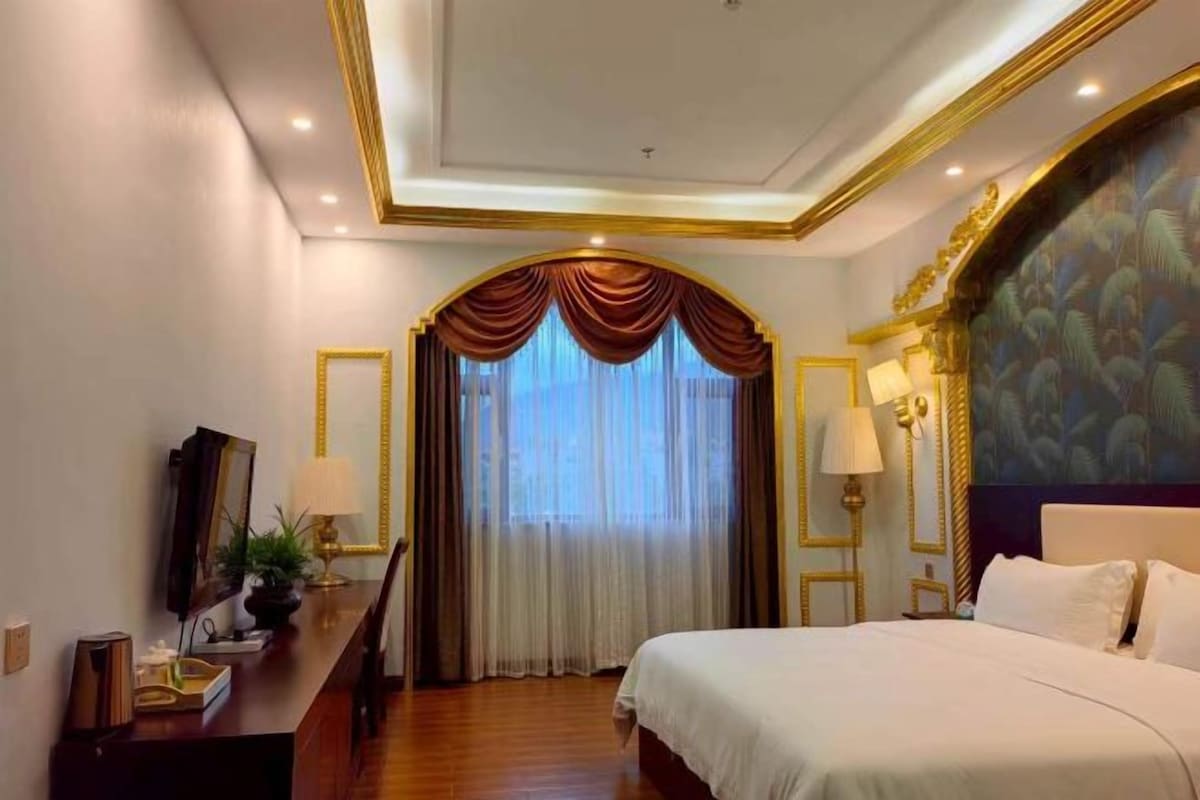 A hotel room with a large bed, a desk with a lamp, a mirror, and a view through a window with curtains.