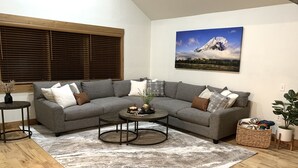 A modern living room with a grey sectional sofa, a round coffee table, and a large painting of a mountain landscape. - Your Montana Home Away From Home (Whitefish)