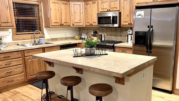 A modern kitchen with wooden cabinets, a stainless steel refrigerator, a microwave, and a central island with stools. - Your Montana Home Away From Home (Whitefish)
