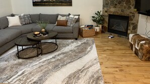 A living room with a gray sectional sofa, a coffee table, a stone fireplace, and a large framed picture of a mountain. - Your Montana Home Away From Home (Whitefish)