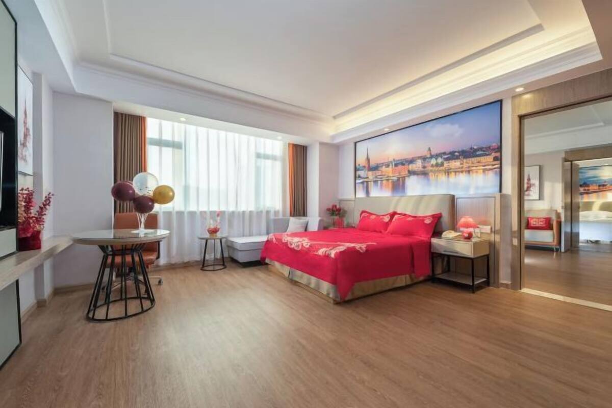 A hotel room with a bed, a desk, a chair, a small table, and a large window.