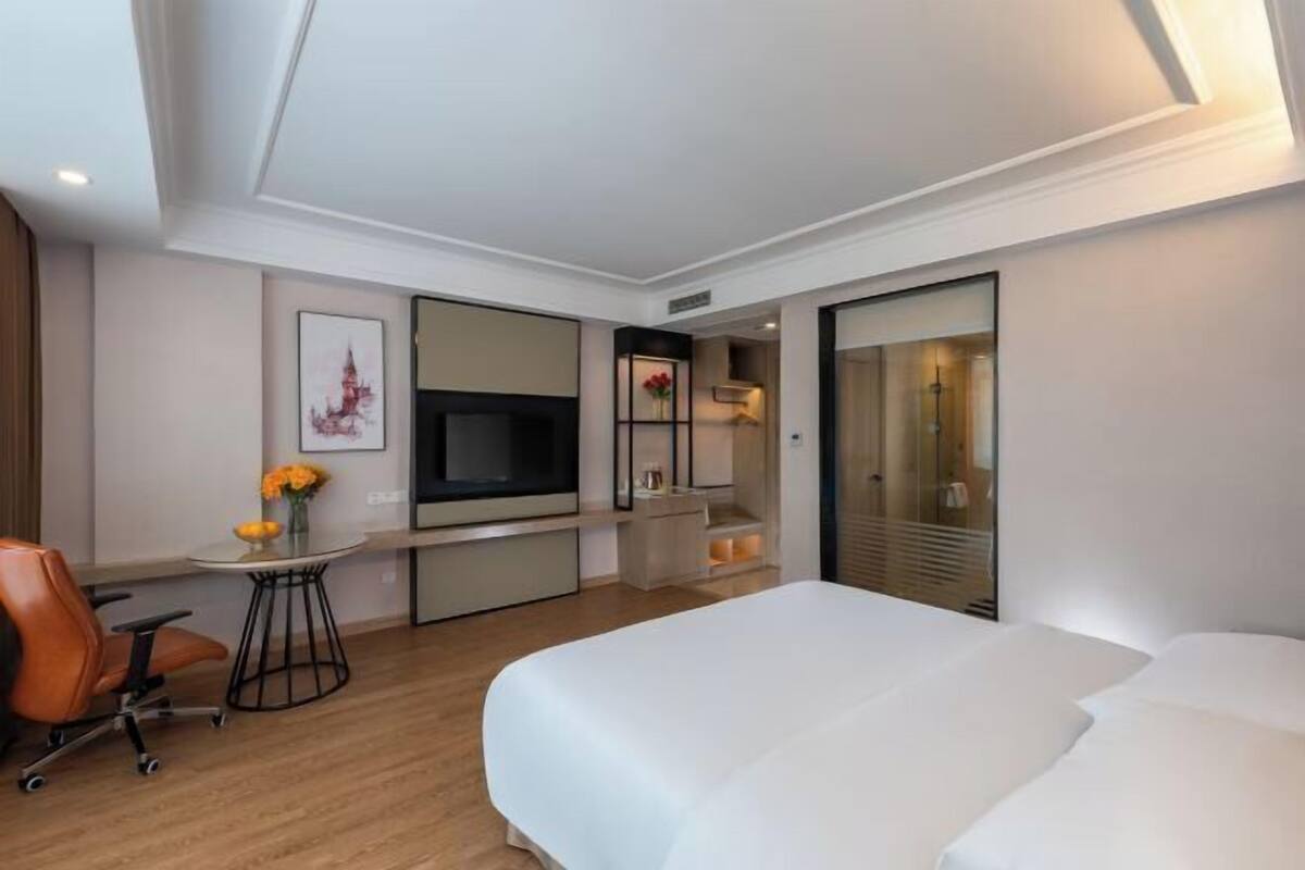 A modern hotel room with a bed, desk, chair, TV, and a bathroom with a glass door.