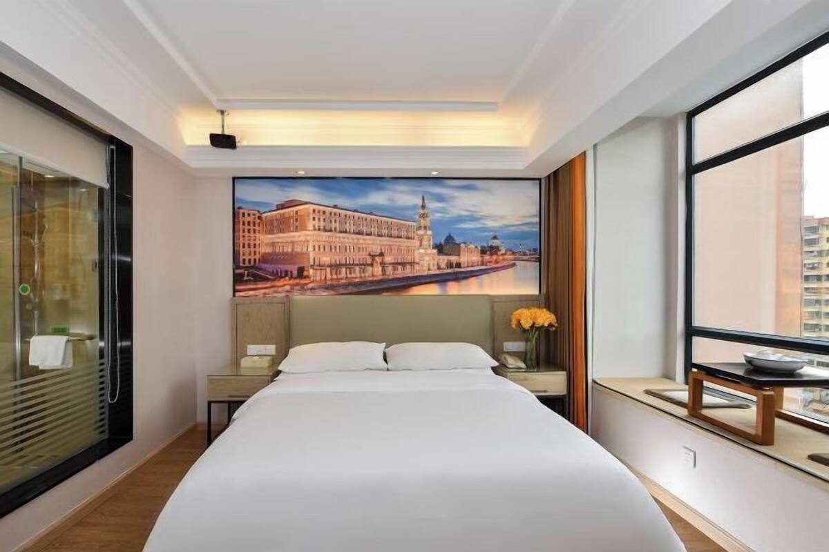 A hotel room with a large bed, a nightstand, a mirror, a window with a view of a building, and a painting of a cityscape.