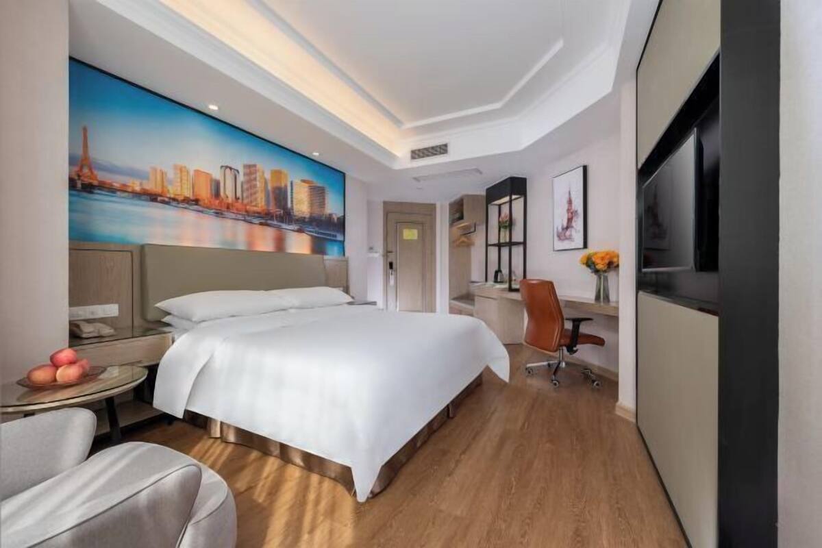 A hotel room with a bed, a desk, a chair, a TV, and a painting of a cityscape.
