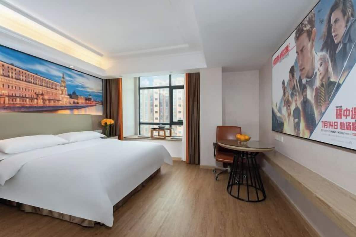 A hotel room with a large bed, a desk with a chair, a window with a view of buildings, and a poster on the wall.