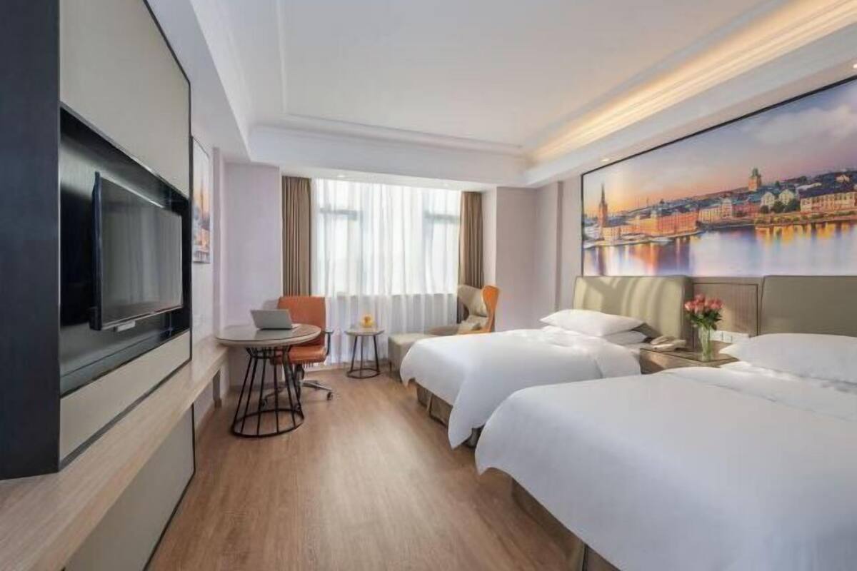 A hotel room with two beds, a TV, a small table with a chair, and a painting of a cityscape.