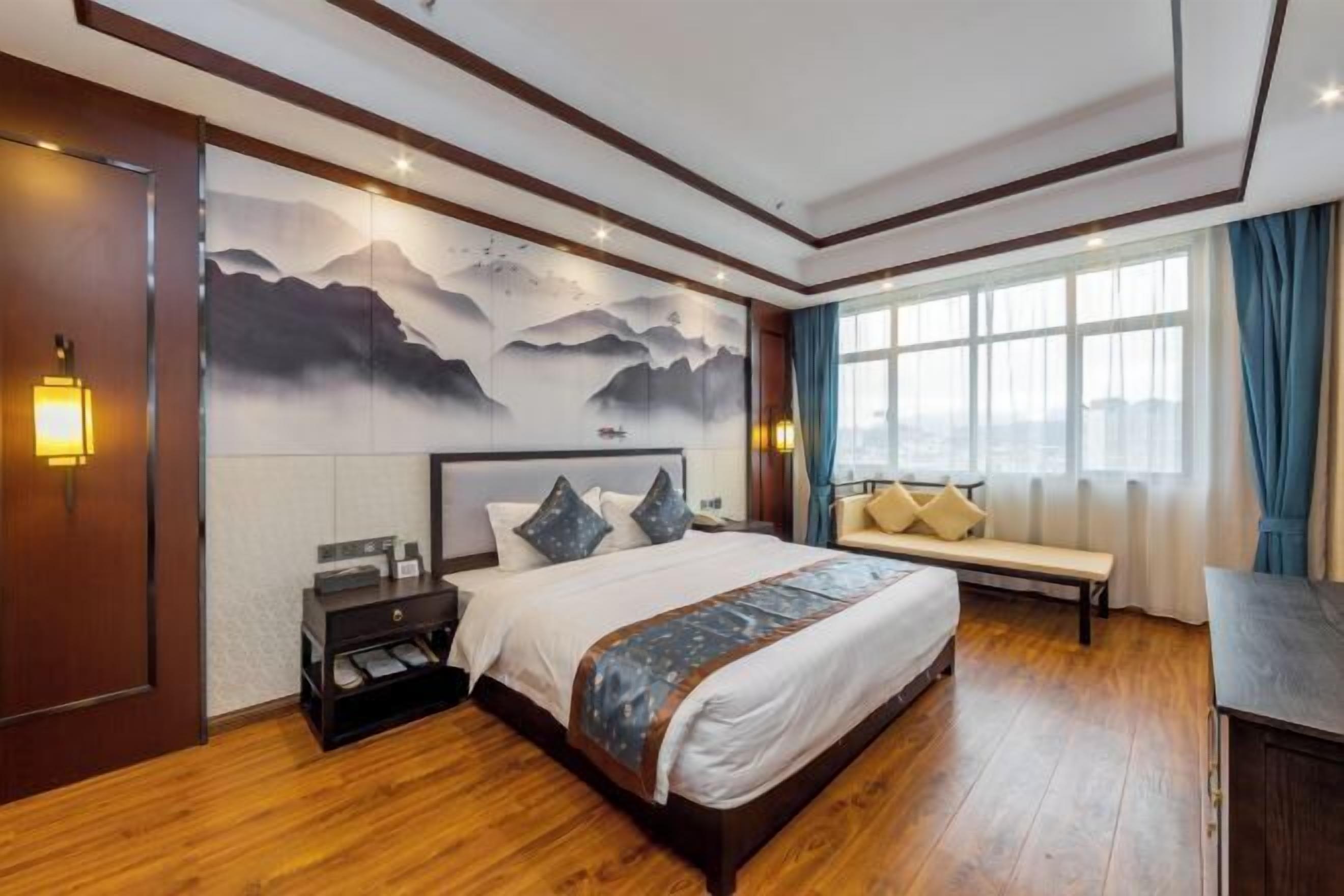Une chambre d’hôtel avec un plancher en bois, un lit avec des oreillers, un banc, une table de chevet, un mur orné d’un tableau représentant une montagne et une fenêtre avec des rideaux.