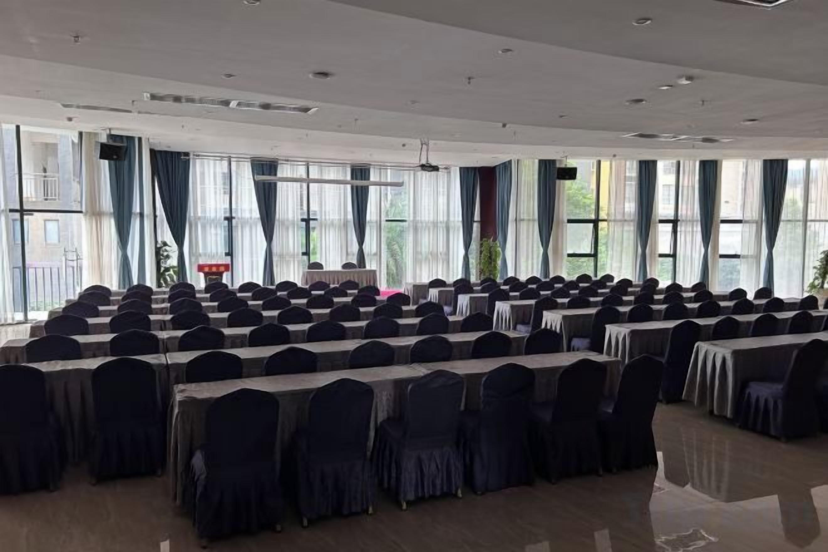 Un salón de conferencias con filas de mesas y sillas, ventanas amplias y un escenario.